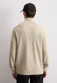 Beige gebreide trui met een hoge kraag, gestructureerde stof en geribbelde manchetten. Zwarte pet achterstevoren gedragen. Minimalistisch ontwerp, geen patronen.