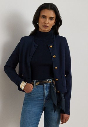 Donna che indossa un cardigan blu navy abbottonato, dolcevita nero a coste, jeans blu, cintura foulard con fantasia e braccialetti dorati, in piedi davanti a uno sfondo uniforme.