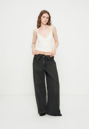 Młoda kobieta poprawiająca luźne czarne szerokie jeansy, ubrana w białą koszulkę na ramiączkach z koronką i czarne buty, stojąca na tle jednolitej białej ściany.