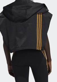 Černá zkrácená bunda s kapucí, na zadní straně se třemi vertikálními zlatými pruhy. Hladká textura s uvolněným střihem.