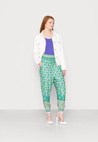 Calças verdes estampadas com cintura ajustável por cordão, combinadas com uma blusa roxa e uma jaqueta de denim branca. Modelo usa saltos brancos.