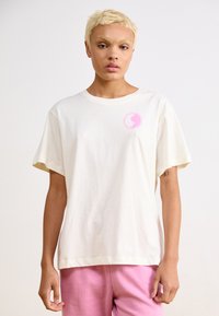 Kortærmet creme t-shirt i bomuld med et lille pink yin-yang grafik på brystet. Løs pasform, standard rund halsudskæring.