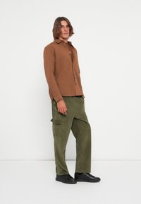 Bruine button-up shirt met lange mouwen, gecombineerd met olijfgroene corduroy cargobroek. Model draagt zwarte sneakers en staat voor een witte achtergrond.