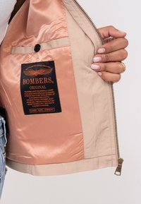 Doublure de veste bomber beige avec une finition satinée lisse. Présente une étiquette noire avec un texte orange, indiquant la marque et des informations supplémentaires.