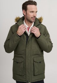 Chaqueta aislante de color verde oliva con capucha forrada de piel, que cuenta con múltiples bolsillos delanteros y cierre de cremallera. Tejido texturizado con costuras en contraste.