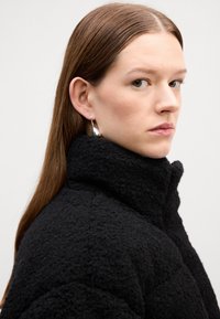 Svart teksturert jakke med høy krage og synlige sømdetaljer. Modellen har på seg sølvfargede hoop-øreringer, langt brunt hår, nøytral bakgrunn.