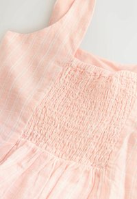 Robe sans manches rose clair avec des rayures verticales blanches et un corsage smocké sur un fond uni.