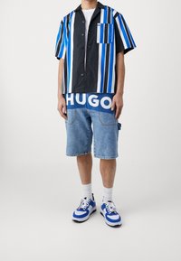 Man in een kortshirt met verticale strepen in blauw, zwart en wit, blauwe shorts met de tekst "HUGO", witte sokken en blauwe en witte sneakers.