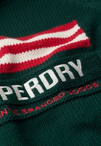 Tissu en tricot vert avec un motif rayé rouge et blanc. Texte brodé "SUPERDRY" en blanc et "AUTHENTIC BRANDED GOODS" en rouge.