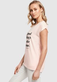 Lys rosa bomulds t-shirt med korte, rullede ærmer, der har sort tekst, som siger "gode ting tager tid" i en moderne skrifttype.