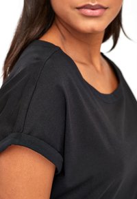 Camiseta de algodón negra con un escote redondo, mangas cortas enrolladas y una textura suave. Primer plano del detalle en el hombro y el escote.