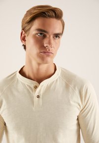 Svetlo bež dolga majica z dolgimi rokavi in henley ovratnikom, dvema lesenima gumboma in raglan rokavi. Mehka tekstura in minimalističen dizajn.