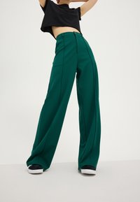 Pantalon vert à jambes larges en tissu lisse, avec une taille haute et des détails le long des coutures latérales, porté avec des baskets noires.