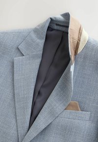 Veste blazer gris clair texturée avec doublure intérieure noire et détail de col beige, montrant un mouchoir de poche beige dans la poche poitrine.