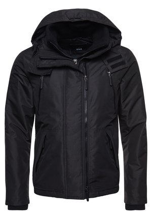 Veste noire à capuche avec fermeture éclair frontale, poches poitrine et latérales zippées, manches à poignets et rabat de capuche ajustable avec velcro.
