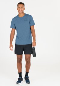 Sininen urheilupaita, jossa on painettuna "ENDURANCE", yhdistettynä mustiin shortseihin. Pitelee mustaa vesipulloa ja jalassa siniset lenkkarit.