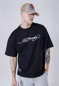 Black cotton t-shirt with white embroidered "Ed Hardy" script. The model wears a beige cap featuring a colorful skull design.