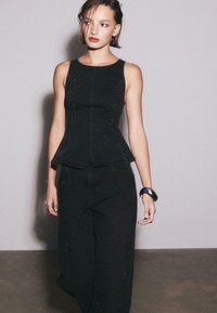 Czarny top bez rękawów z baskinką, zestawiony z czarnymi szerokimi spodniami jeansowymi. Materiał ma strukturę z subtelnymi zdobieniami. Dodatki obejmują gruby czarny bransoletkę.