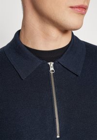 Cămașă tricotată de culoare bleumarin, cu fermoar și guler clasic. Țesătura pare moale, oferind un aspect texturat.