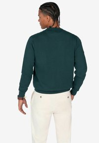 Dunkelgrüner Sweatshirt mit Rundhalsausschnitt und gerippten Bündchen, getragen über hellen Hosen, mit minimalistischem Design und ohne sichtbare Muster.