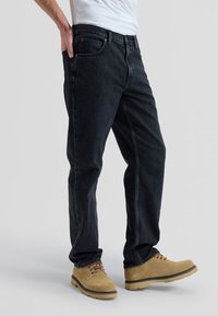 Man draagt zwarte jeans en beige veterlaarzen, staand tegen een eenvoudige lichte achtergrond.