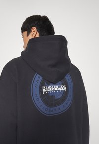 Sudadera con capucha negra con un gráfico circular azul en la parte trasera que muestra "NAPAPIJRI geographic." El material parece suave con un acabado texturizado.