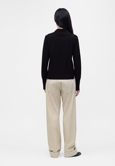 Femme aux longs cheveux foncés portant un haut noir à manches longues et un pantalon beige ample, debout de dos sur un fond blanc uni.
