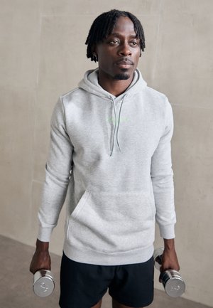 Gray sweatshirt with a hood, featuring a front pocket and green logo. Model holds dumbbells, wearing black shorts. Smooth texture.