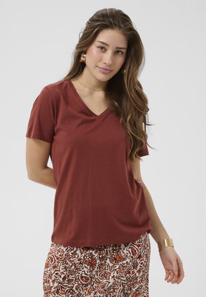 Donna con capelli lunghi che indossa una maglietta a V di colore ruggine e una gonna bianca con motivi floreali arancioni, in piedi con una mano dietro la schiena.