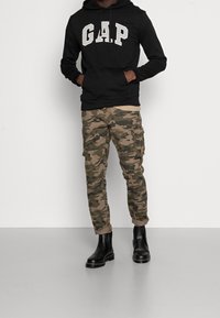 Felpa nera con cappuccio e logo "GAP" bianco, abbinata a pantaloni cargo camouflage e stivali neri alla caviglia, in piedi su uno sfondo neutro.