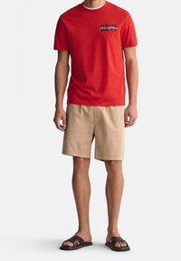 Rotes Baumwoll-T-Shirt mit Grafikdruck auf der linken Brust, kombiniert mit beigen Shorts und braunen Sandalen. Lässiges und bequemes Outfit.