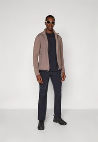 Šviesiai ruda užtraukiama striukė dėvima ant tamsios marškinėlių, derinama su tamsiomis kelnėmis ir juodais batais. Striukė turi tekstūruotą dizainą ir neturi matomų logotipų.