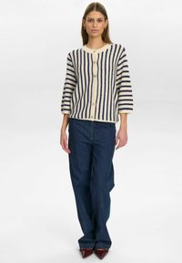 Strickjacke mit vertikalen marineblauen und cremefarbenen Streifen, runder Ausschnitt, Dreiviertelärmel und vorderer Knopfverschluss. Kombiniert mit dunklen Jeans.