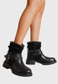 Jambes portant des bottes en cuir noires mi-mollet avec des sangles à boucle, fermeture éclair latérale et tige en daim texturé sur un fond gris clair.