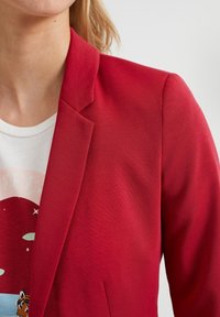 Röd blazer med en jämn textur, slagkrage och skräddarsydd passform, matchad med en vit grafisk t-shirt med en färgglad design.