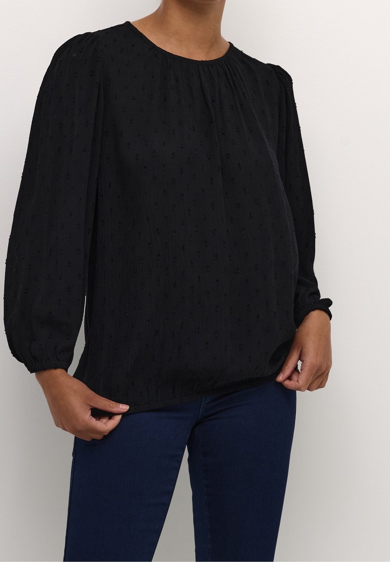 Femme portant un chemisier noir texturé à manches longues avec un col froncé et un pantalon bleu marine, les mains tenant l'ourlet du chemisier.