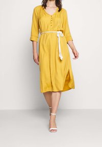 Żółta sukienka do kolan z rękawami trzyczwartowymi, z zapięciem na guziki z przodu oraz białym plecionym paskiem akcentującym talię. Widoczny detal z rozcięciem po boku.