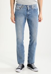 Jeans de mezclilla de color azul claro con corte recto, cinco bolsillos y detalles en hardware de cobre. Llevados con zapatillas negras de caña alta.