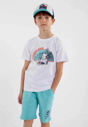 T-shirt en coton blanc avec un graphisme "Timberland" rouge et turquoise. Shorts aqua avec poches latérales et un écusson logo noir. Casquette ajustable avec un design de montagne.