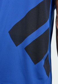 Top deportivo azul con textura de malla que presenta grandes formas geométricas negras y acentos. El diseño incluye paneles azules en contraste.