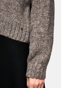 Un pull en tricot marron avec un ourlet et des poignets côtelés, texture douce, présentant un petit logo. Porté par-dessus un vêtement noir.
