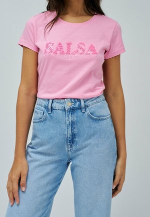 Kobieta w różowym dopasowanym t-shircie z napisem „SALSA JEANS” i jasnoniebieskich, wysokich dżinsach na jednolitym tle.