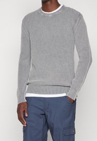 Harmaa neulepaita, jossa on resorit kauluksessa ja hihoissa. Helmassa vaakasuora raidallinen kuvio. Päällä valkoinen paita.