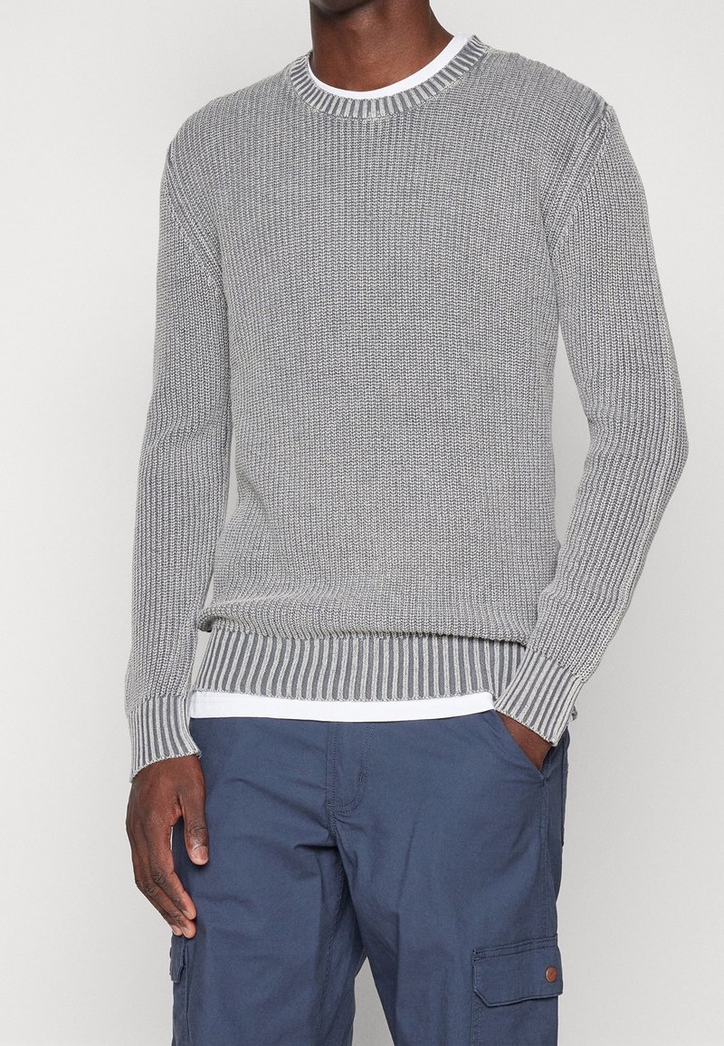 Harmaa neulepaita, jossa on resorit kauluksessa ja hihoissa. Helmassa vaakasuora raidallinen kuvio. Päällä valkoinen paita.