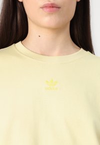 Svetložlté bavlnené tričko s rebrovaným výstrihom a malým žltým vyšívaným logom Adidas na hrudi. Hladká textúra.
