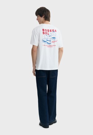 Młody mężczyzna w białej koszulce z czerwono-niebieskim nadrukiem i ilustracjami jedzenia na plecach, w ciemnoniebieskich spodniach i czarnych butach.