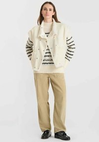 Ivoor vest met schapenvacht kraag en drukknoopjes, gedragen over een zwart-wit gestreepte trui, gecombineerd met beige corduroy broek en zwarte schoenen.