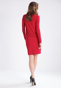 Robe rouge longueur genou avec manches longues, fabriquée en tissu lisse. Le dos présente une coupe décontractée, associée à des chaussures à talons hauts noires.