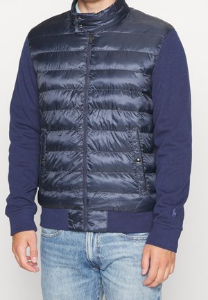 Homme portant une veste matelassée bleu marine avec fermeture éclair sur une chemise bleu marine à manches longues et un jean, debout devant un fond clair uni.