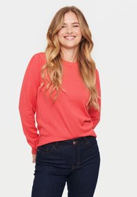 Pulover lung cu mâneci lungi de culoare corai, cu decolteu rotund și umeri strânși, asortat cu blugi bleumarin, cu închidere cu button și buzunare laterale.